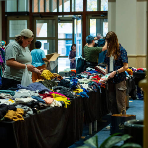 Rows of tables and clothing racks with brightly colored clothes fill the frame, as several attendees peruse the items.